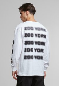 White long-sleeve shirt featuring a black spray-painted "ZOO YORK" text pattern on the back and sleeves, with a relaxed fit and cotton material.