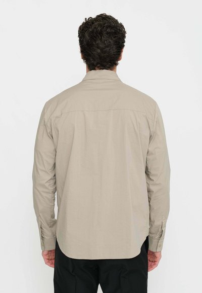 Camicia beige a maniche lunghe con colletto standard, cucitura sulla spalla posteriore e polsini con bottoni. Realizzata in tessuto leggero, presenta un orlo curvo.