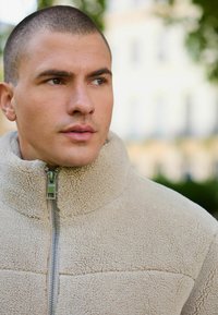 Jachetă din fleece bej cu guler înalt, suprafață texturată și fermoar frontal. Prezintă un capăt de fermoar argintiu. Fundalul este ușor neclar.