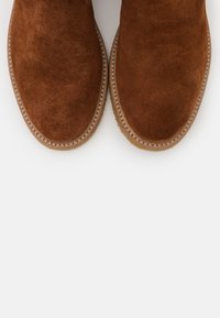 Brown suede ankle boots with a rounded toe and stitched detailing. The sole features a light brown rubber material.