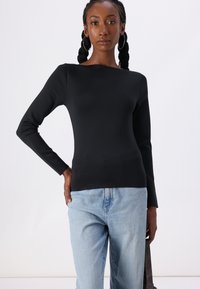Black long-sleeve top with a boat neckline, made of stretch fabric. Paired with light blue denim jeans, model holds a bag.