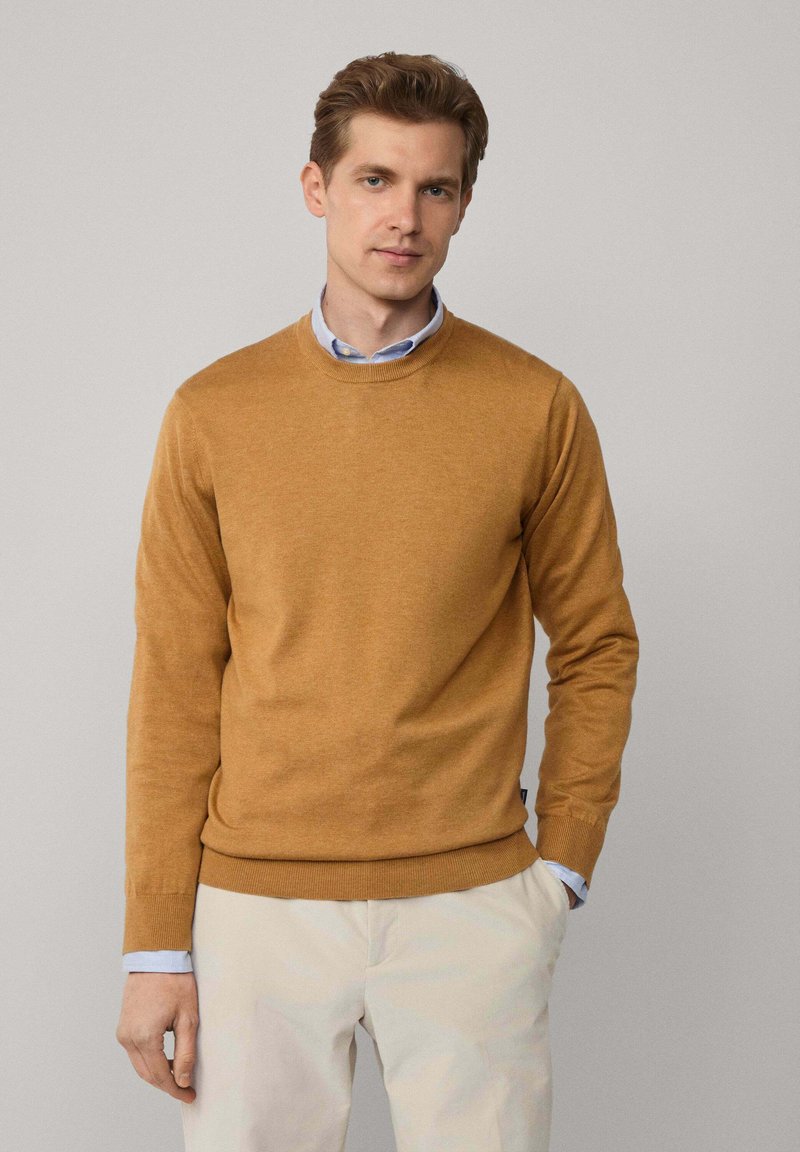 Musztardowożółty sweter z okrągłym dekoltem wykonany z miękkiej dzianiny, z długimi rękawami i żebrowanym wykończeniem, zestawiony z jasnymi spodniami.