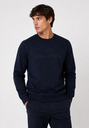 Homme portant un sweat-shirt bleu marine avec « Paul & Shark » et un logo de requin, mains dans les poches, regardant vers la gauche sur un fond clair uni.