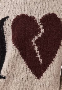 Πλεκτό πουλόβερ με κρεμ βάση και σχέδιο σπασμένης καρδιάς σε μπορντό χρώμα με μαύρες λεπτομέρειες, που αναδεικνύει υφή απαλή και αφράτη.