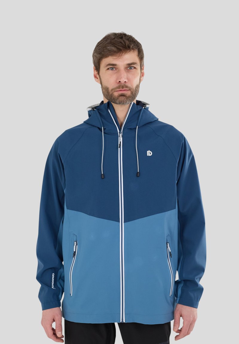 Mies, joka käyttää kaksiväristä sinistä hupullista takkia, jossa on etuvetoketju ja vetoketjulliset taskut, seisoo yksinkertaisen vaaleanharmaan taustan edessä.