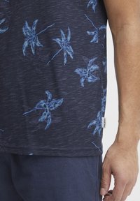 Tamsiai mėlyna marškinė su tekstūruota medžiaga, dekoruota šviesiai mėlynu tropiniu palmės lapų raštu. Ant šono matomas etiketė.