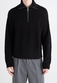 Schwarzer Pullover mit Zip-Kragen und strukturiertem Rippstrickdesign sowie langen Ärmeln. Trägt man über hellgrauen, locker geschnittenen Hosen.