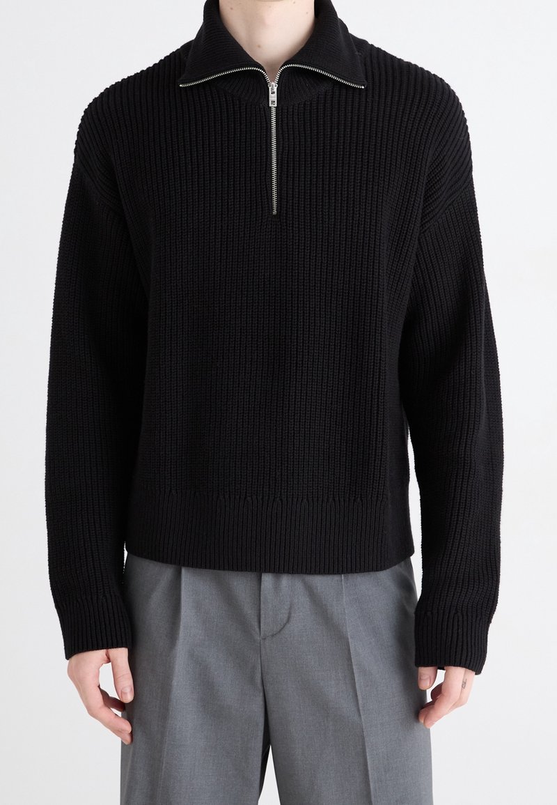 Schwarzer Pullover mit Zip-Kragen und strukturiertem Rippstrickdesign sowie langen Ärmeln. Trägt man über hellgrauen, locker geschnittenen Hosen.