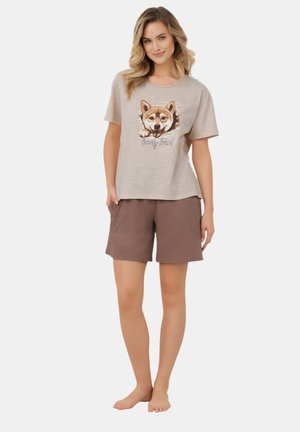 Kobieta stojąca boso, ubrana w beżowy t-shirt z wizerunkiem psa i napisem „family friend”, w połączeniu z brązowymi szortami o długości do kolan.