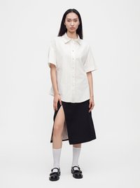 Jeune femme portant une chemise blanche à manches courtes boutonnée et une jupe midi noire avec une fente haute sur le côté, associée à des chaussettes blanches et des chaussures noires.