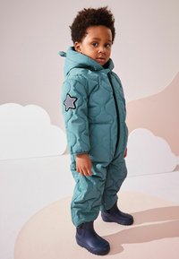 Combinaison de neige matelassée teal avec capuche, ornée d'une patch en étoile sur le bras gauche, fermeture éclair, accompagnée de bottes en caoutchouc bleues.