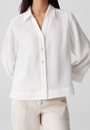 Woman wearing a loose white textured button-up shirt with a collar and long sleeves, paired with beige pants.