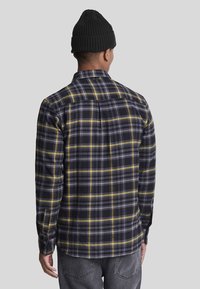 Camisa de cuadros en negro, gris y amarillo con mangas largas y puños con botones. Cuenta con canesú trasero y costuras en los hombros, fabricada con tela suave.