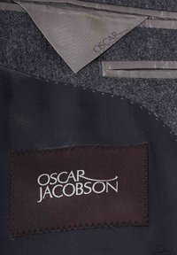 Szary materiał wełniany z gładką czarną satynową podszewką oraz metką z napisem "Oscar Jacobson" na białym tle na ciemnym tle.