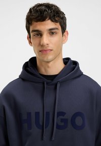 Giovane uomo con capelli corti e ricci che indossa un hoodie blu navy con "HUGO" in grandi lettere sul davanti, su uno sfondo semplice.