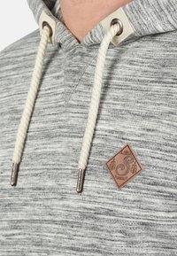 Gray heathered pullover hoodie with a ribbed texture, featuring a drawstring closure, metal eyelets, and a brown leather logo patch.