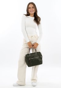 Olivově zelená kabelka z texturované látky, s přední kapsou na zip a dvojitými ručkami. Modelka má na sobě bílý top a světlé kalhoty.