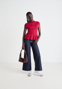 Kvinne som har på seg en rød ermeløs peplum-top, vide mørkeblå denimjeans, hvite joggesko, solbriller, og holder en mørkebrun håndveske foran en enkel bakgrunn.