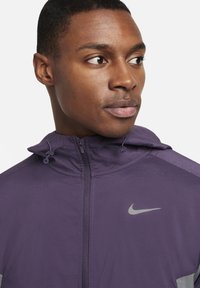 Chaqueta Nike púrpura con cierre de cremallera, capucha y tejido texturizado. Presenta acentos grises y logo en el pecho. Vista en primer plano del diseño.