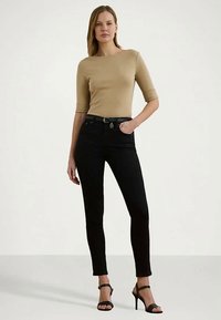 Ženska stoji z roko v žepu, oblečena v bež oprijet zgornji del, črne oprijete kavbojke in črne sandalke z visokim petkom na enotnem ozadju.