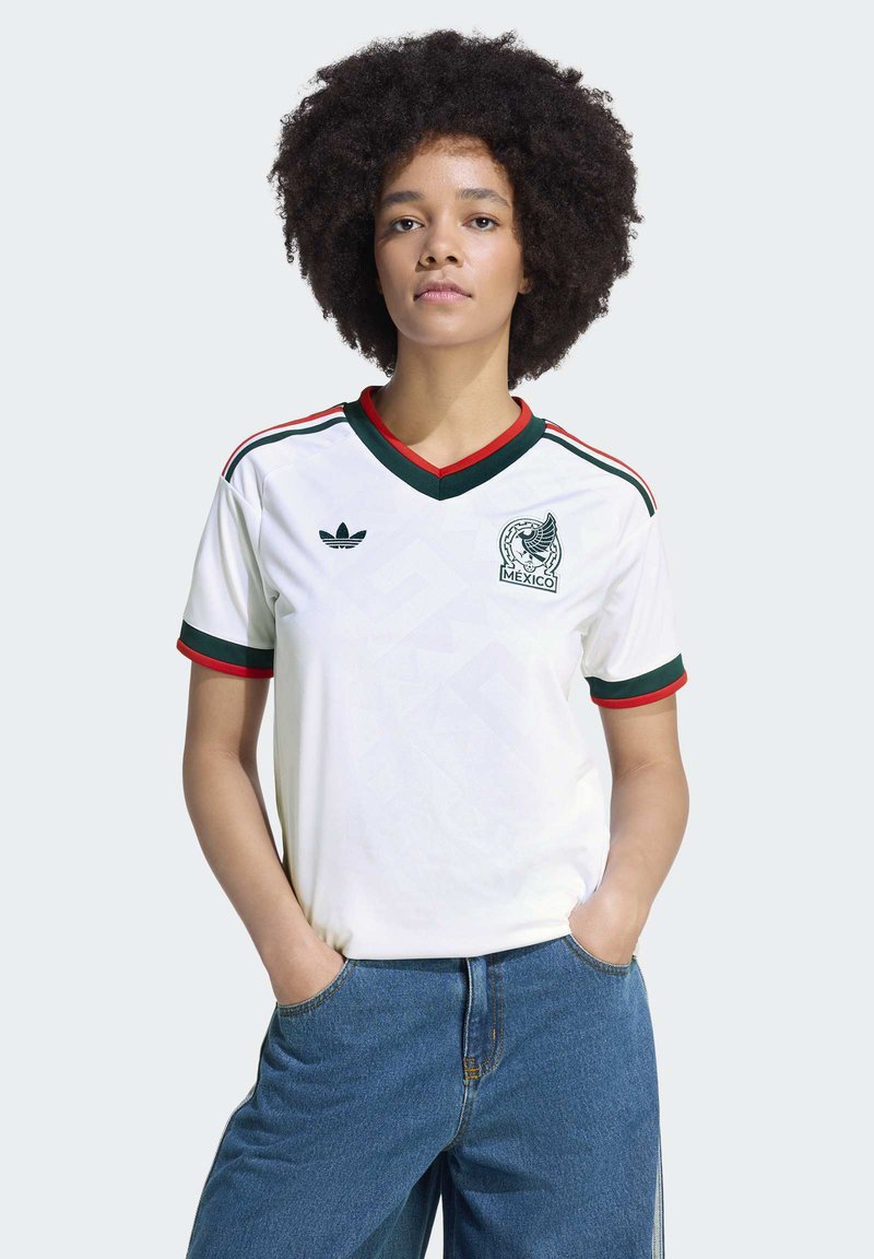Jeune femme avec une coiffure afro portant un maillot de football du Mexique blanc avec des bordures vertes et rouges, debout les mains dans les poches d'un jean bleu.