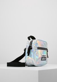Bolso de hombro holográfico con cierre de cremallera, correa ajustable negra y dos destacados logotipos de Adidas en negro en el frente.