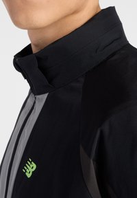 Zwarte jas met grijze accenten, hoge kraag, ritssluiting aan de voorkant en een klein groen logo aan de zijkant. Gladde textuur en een aansluitend ontwerp.