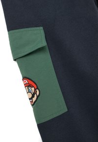 Donker marineblauwe stof met een groene zak waarin een geborduurde patch van het gezicht van een cartoonfiguur met een rode hoed is verwerkt.