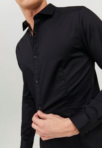 Černá košile na knoflíky s hladkou texturou, špičatým límečkem a dlouhými rukávy. Má přední knoflíky a detaily přizpůsobeného střihu.