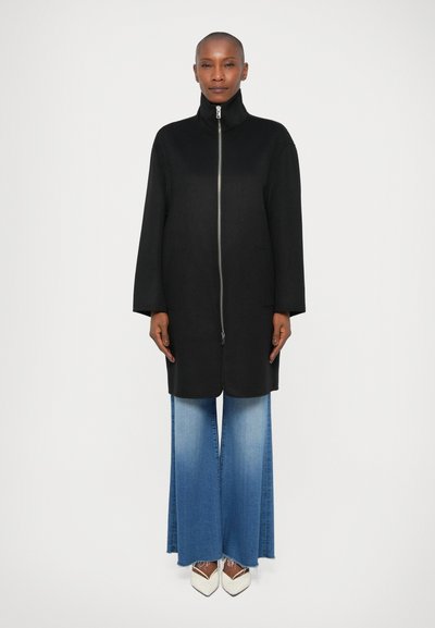 Manteau noir avec un col montant et une fermeture éclair sur toute la longueur, associé à un pantalon en denim bleu clair évasé et des chaussures blanches à bout pointu.
