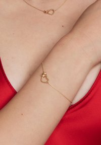 Złota bransoletka z łańcuszka z sercowym zapięciem. Łańcuszek jest delikatny z teksturowanymi akcentami. Kolor skóry kontrastuje z czerwoną odzieżą.