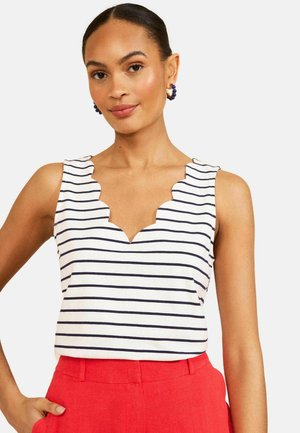 Kobieta z włosami zaczesanymi do tyłu, ubrana w biały, bezrękawnik z granatowymi paskami i karbowanym dekoltem, w połączeniu z wysokim stanem czerwonych spodni.