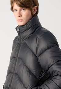 Casaco puffer preto com gola alta, costura diagonal e fecho à frente, feito de um material liso e brilhante.