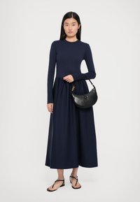 Jūras karafles garām piedurknēm kleita ar pieguļošu augšdaļu un plūstošu svārku. Melna ādas rokassoma ar izliekta forma. Modelis valkā plakano sandali.