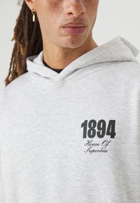 Hoodie gris clair avec une coupe décontractée, ornée d'un texte imprimé noir "1894" et "House Of Superbia" sur la poitrine, en tissu doux.