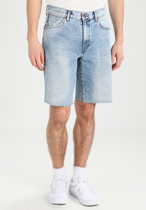 Lichtblauwe denim shorts met een rechte snit, rafelige onderrand, vijf zakken en koperen details, gecombineerd met witte sneakers en sokken.