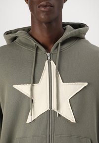 Felpa con zip di colore verde oliva, con un grande design di una stella bianca sul davanti, caratterizzata da una texture morbida e cappuccio con cordini. Dettaglio della zip argentata.