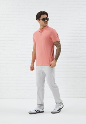 Camisa de polo rosa con cuello, combinada con pantalones gris claro. Zapatillas deportivas con detalles en negro. El modelo lleva gafas de sol y tiene tatuajes.