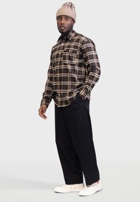 Camisa xadrez castanha de botão com uma bainha arredondada, combinada com calças pretas e ténis brancos, rematada com um gorro de malha bege.