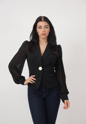 Mujer con cabello largo y oscuro, vistiendo un blazer negro con mangas abullonadas de tela transparente y un botón decorativo, combinado con vaqueros azul oscuro.