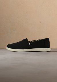 Chaussure en toile noire à enfiler avec semelle blanche et petite étiquette latérale sur fond neutre.