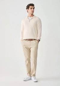 Mężczyzna stojący z rękoma w kieszeniach, ubrany w jasnobeżowy sweter, pasujące spodnie, białe trampki, brązowy pasek oraz zegarek na tle jednolitym.
