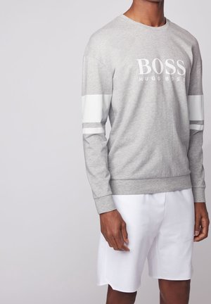Mand iført lysegrå langærmet sweatshirt med hvide striber på ærmerne og "BOSS HUGO BOSS" logo, kombineret med hvide shorts.