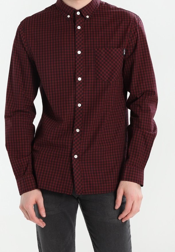 Homme portant une chemise à carreaux rouge foncé et noire à manches longues avec une poche poitrine et un pantalon noir.