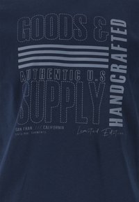 T-paita tummansinisenä, jossa on valkoista graafista tekstiä. Tekstissä lukee "GOODS &", "AUTHENTIC U.S. SUPPLY" ja "HANDCRAFTED" viivayksityiskohtineen.