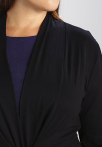 Top avvolgente nero a maniche lunghe con tessuto drappeggiato, caratterizzato da una texture morbida. Sotto, è parzialmente visibile un top fitted viola.