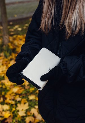 White leather wallet with a zippered closure, held in black gloved hands, set against a backdrop of yellow autumn leaves.