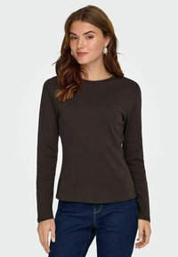Langsleeve bruin top van zacht materiaal met een ronde halslijn en naaddetails, gedragen met donkerblauwe jeans. Eenvoudig en getailleerd ontwerp.
