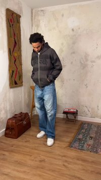 Νέος άνδρας με μαύρη μπλούζα με κουκούλα και τζιν κοιτάζει προς τα κάτω, όρθιος σε ένα δωμάτιο με φθαρμένους τοίχους, ξύλινο πάτωμα, καφέ τσάντα, καρέκλα, μικρό τραπέζι και χαλί με σχέδια.