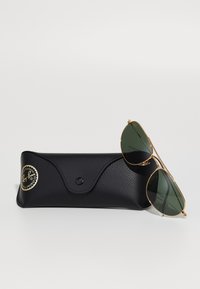 Occhiali da sole Ray-Ban aviator con lenti verdi, montatura in metallo dorato e astuccio in pelle nera testurizzata con logo circolare e chiusura magnetica.